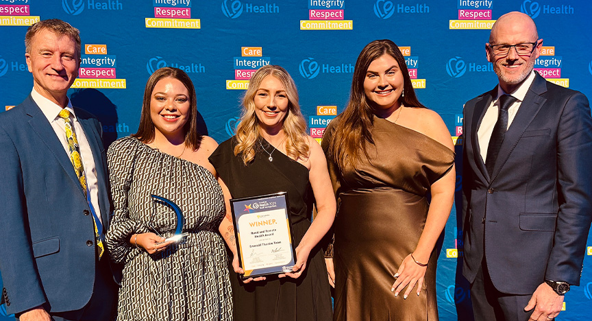 Sponsor Central Queensland University Vice-Chancellor Professor Nick Klomp, and Executive Director of Nursing and Midwifery Andy Carter congratulate members of Emerald Hospital’s Operating Theatre team Danae Louie, Courtney Saiat and Rebecca Carver on their win in the Rural and Remote Health Award. Healthcare workers wearing cocktail attire stand in front of a royal blue media wall printed with Central Queensland Health values at an awards presentation evening at a conference centre.