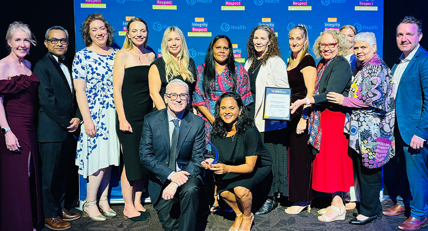 Winner of the Health Equity Award, members of the Central Queensland Health Tuberculosis Team. From left: Susie Le Brasse, Professor Gulam Khandaker, Dr Jemma Taylor, Evie Perrins, Leah Hosemann, Tania Roberts, Margaret Charles, Danielle Odorico, Julie Drayton, Jacina Walker (obscured), Betty Doyle, Dr Michael Kirk, and kneeling Dr Mark Stickley and Katrina Ingra. Healthcare workers wearing cocktail attire stand in front of a royal blue media wall printed with Central Queensland Health values at an awards presentation evening at a conference centre.