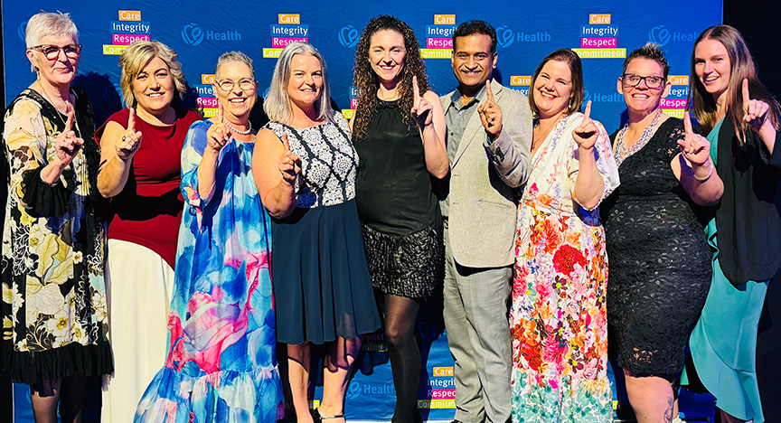 Healthcare workers wearing cocktail attire stand in front of a royal blue media wall printed with Central Queensland Health values at an awards presentation evening at a conference centre.