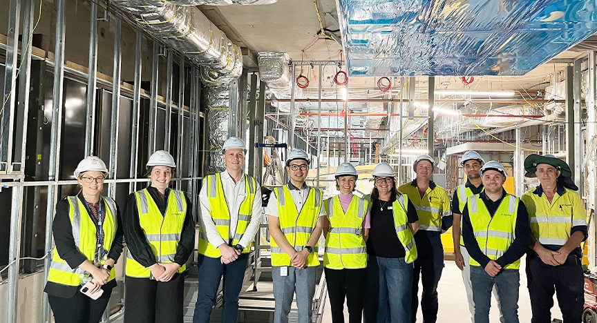 Hospital staff and construction staff all wear hi-vis yellow safety vests and white hard hats standing in a line in a construction zone with steel girders, silver venting and a blue-plastic covered piece of technology on the ceiling.
