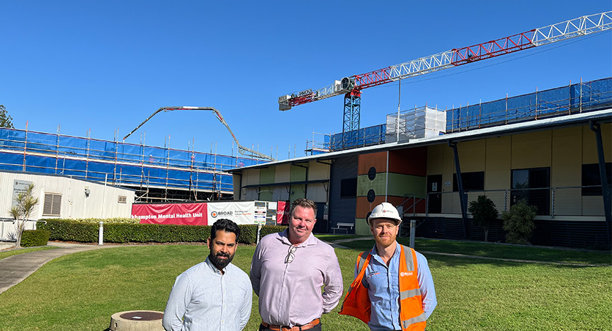 Two men wearing business attire and one wearing high-vis construction gear stand on a grassy area in front of a building site, with a concrete pour and large crane in the background against a blue sky.