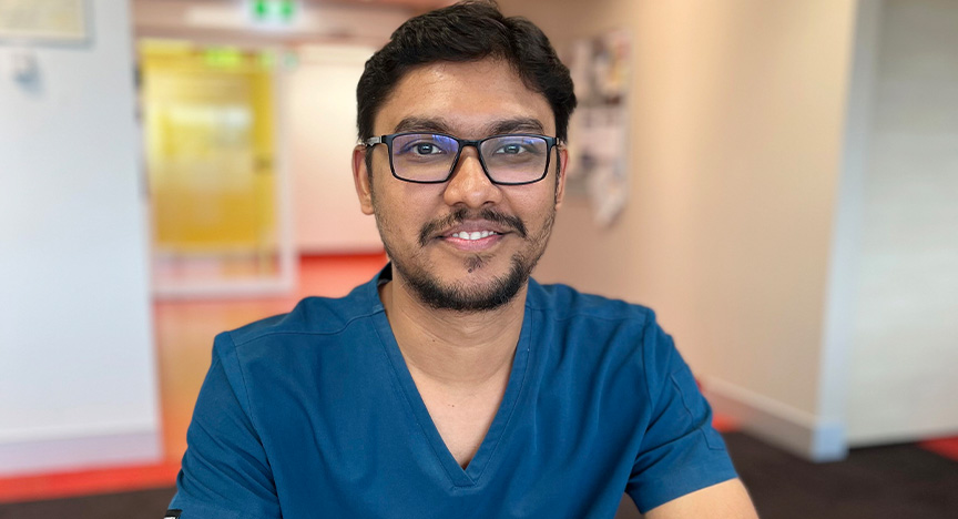 Head and shoulders photo of a doctor wearing a blue scrub top in a hospital meeting room.
