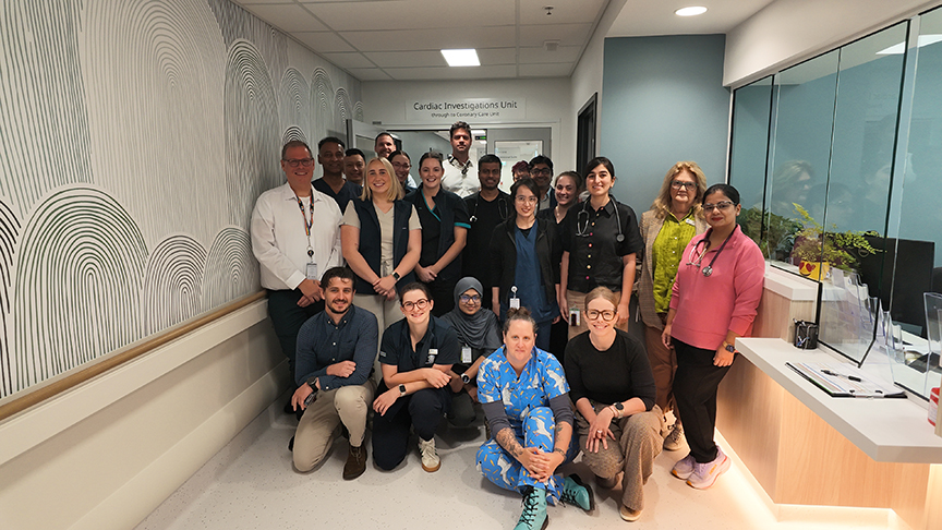 Cardiac Investigations Unit staff standing and sitting in a group smiling at the camera, in the corridor in front of the doorway to the Cardiac Investigations Unit