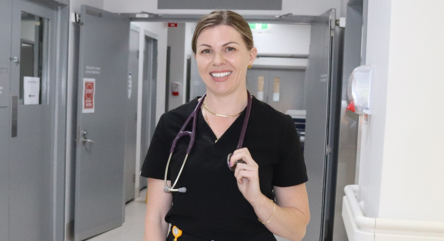 Cassandra wears a black scrub top and holds a stethoscope around her neck with one hand while standing in a hospital corridor.