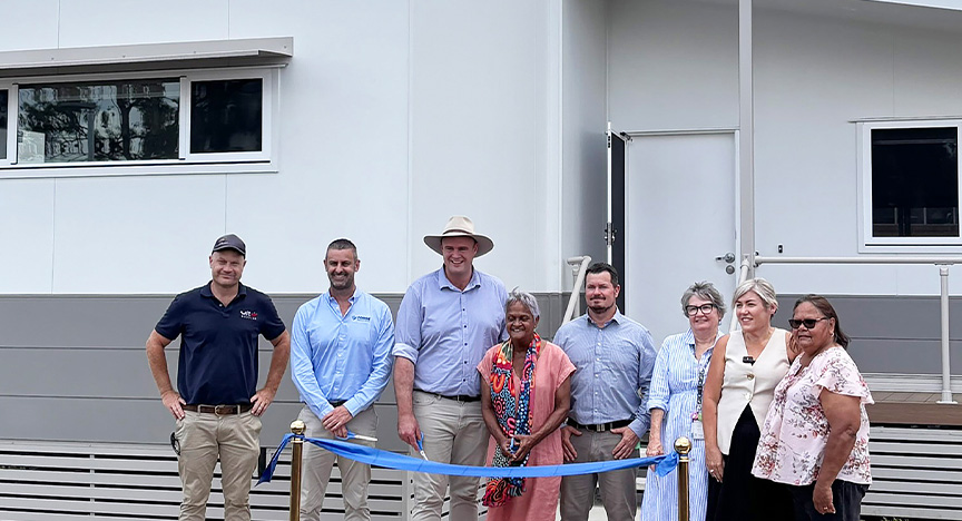 Group of people stand behind two gold bollards with a blue ribbon in front of a new accommodation building on a beautiful sunny day in Biloela.