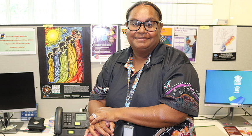A first Nations health worker wearing a black shirt with indigenous print stands in an office with a phone and computers in the background.