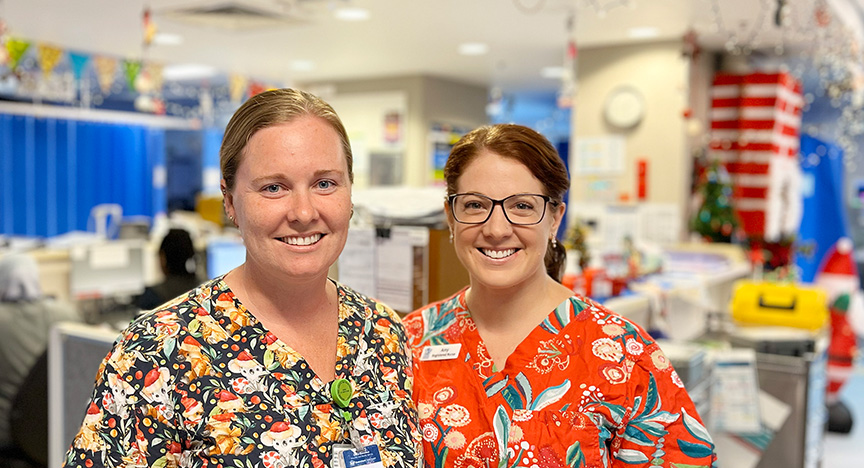 Two registered nurses wearing Christmas-themed scrubs stand in a hospital emergency department adorned with Christmas decorations and twinkling lights, with cubicles screened by blue curtains in the background.