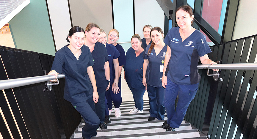 High angle shot of eight of CQ Health's new group of registered nurses, dressed in scrubs, stand together smiling on a stairway at Gladstone Hospital.
