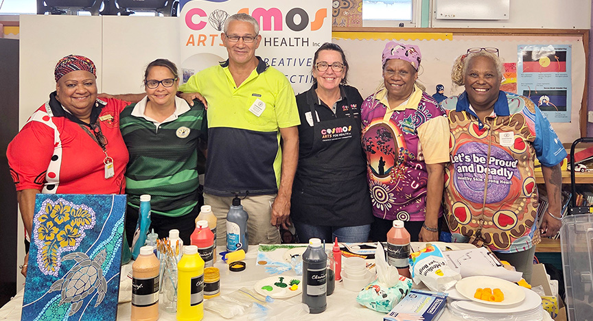 Six smiling adults standing in front of a banner with art supplies on a table in front of them