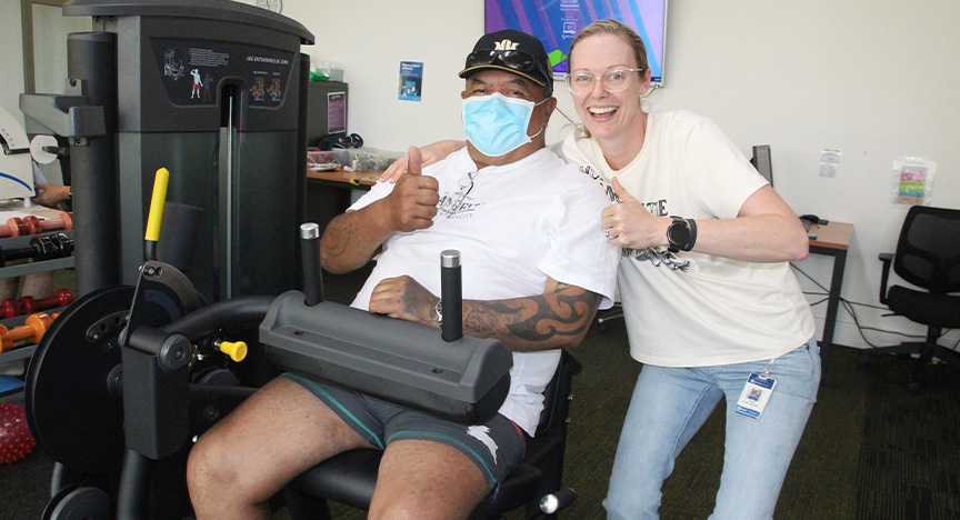 A lung transplant patient wearing a face mask works out on a leg machine in a rehabilitation gymnasium, being hugged by an exercise physiologist, both giving thumbs up.