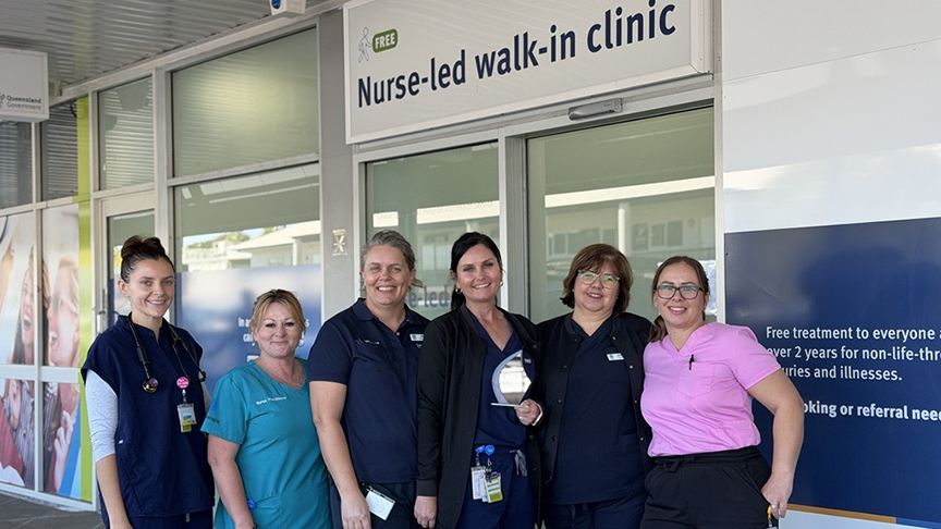 Six staff in front of the Nurse-led walk-in clinic front doors, smiling at camera. One of the staff is holding a staff award that the team won in 2026.