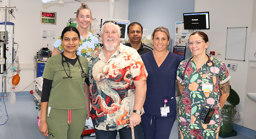 A male patient uses a cane to stand in a treatment bay in an emergency department. He stands with five doctors and nurses wearing scrubs in front of a hospital bed and medical equipment.