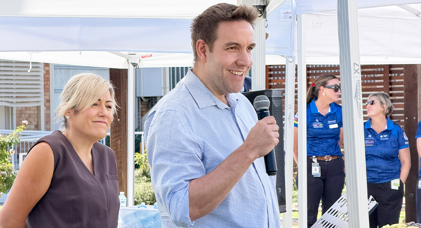 Two executives wearing business attire stand under a marquee, one holding a microphone, in an outdoor area near a hospital, with two health workers wearing royal blue polo shirts standing in the background.