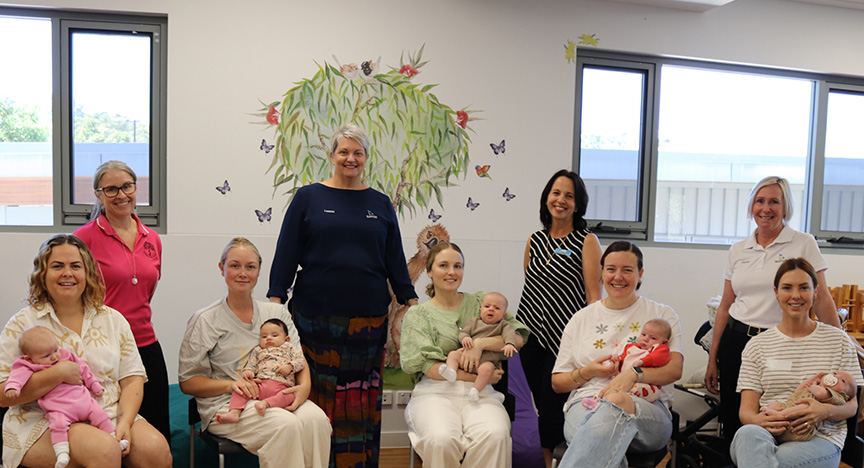 Five mums sit on chairs, smiling and holding their babies, in a bright room decorated with a mural of a tree and butterflies. Four smiling women stand behind the mums and bubs.