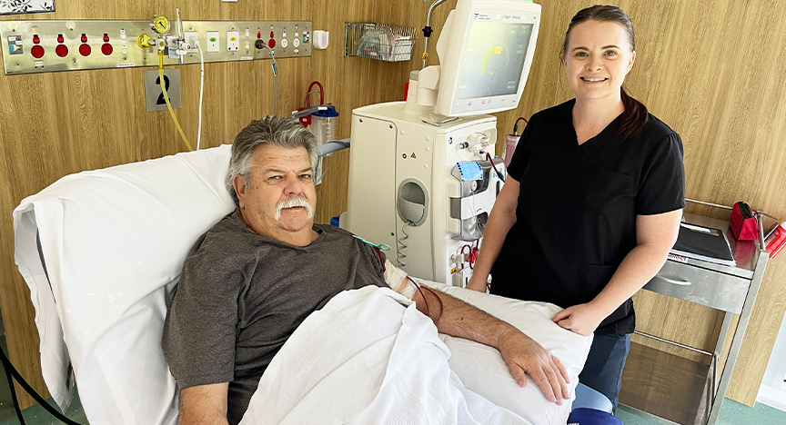 A renal patient sits in a recliner chair with his arm on a pillow while tubes are connected to his arm and a dialysis machine. A renal nurse wearing a black shirt stands behind the patient and in front of the equipment.