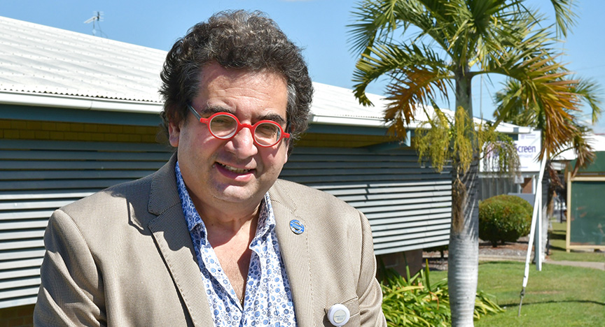 Anaesthetist wears a business shirt and suit coat, red-rimmed circle glasses, stands in front of an administration building with blue sky and green grass.