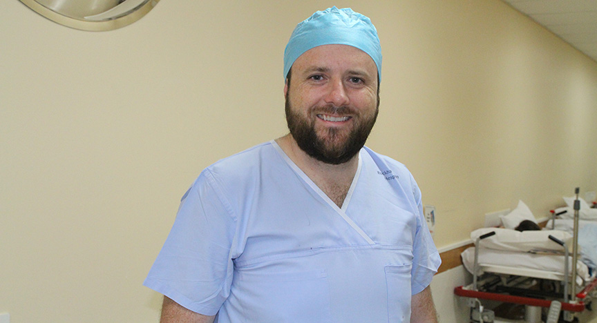 A doctor wearing blue scrubs and surgical cap stands in a hospital operating room with a patient lying on a bed in the background.