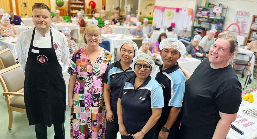 A chef wearing a white uniform and black apron stands with a director of nursing in a colourful dress, three cooks wearing a uniform of navy, white and sky blue sleeves, and an operational services manager wearing a black t-shirt, in a recreation hall at a nursing home, with residents seated at tables wearing party hats in the background.