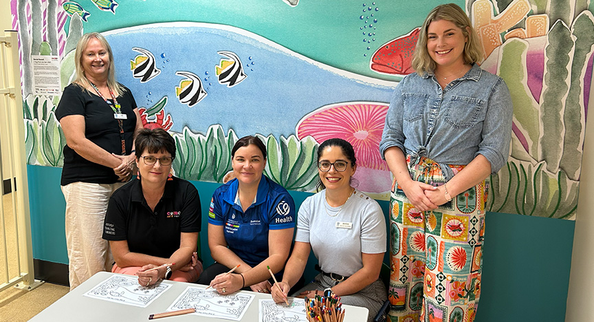 Five women behind a colouring-in table in front of an ocean-themed mural in a hospital building.