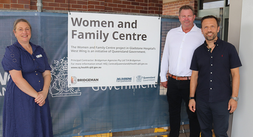 A hospital manager stands with construction staff outside in front of a building with a sign reading “Gladstone Women and Family Centre”.