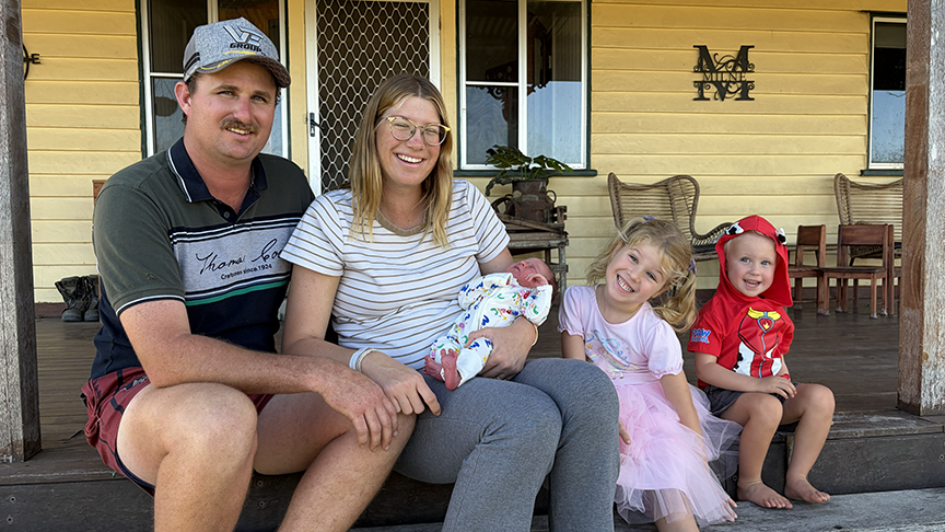 The Milne family - dad, mum holding baby, and young daughter and son, sitting on their stairs on the front porch at home, smiling at camera.