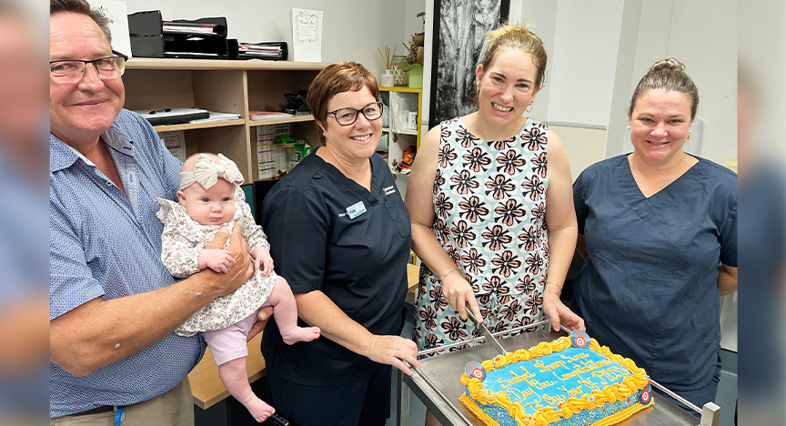 Nursing director holds a baby and three clinical nurse consultants stand behind a birthday cake on a stand in a hospital office