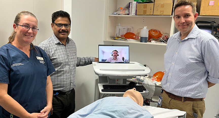Three clinicians stand around a training mannequin covered in a sky blue blanket in a staff simulation training room in a hospital, with a doctor on a mobile unit laptop.