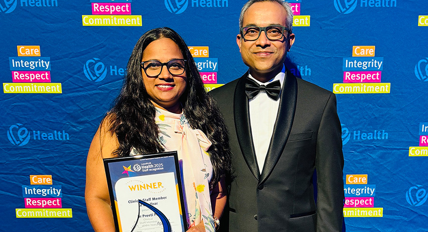 Clinical Staff Member of the Year Dr Preeti Patil is congratulated by Executive Director of Medical Services Professor Gulam Khandaker. Healthcare workers wearing cocktail attire stand in front of a royal blue media wall printed with Central Queensland Health values at an awards presentation evening at a conference centre.