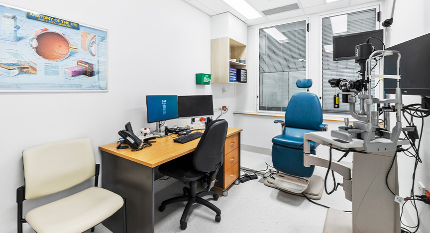 A hospital clinic room featuring a large consultation chair and eye-examining equipment, with a computer desk sitting under an eye chart on a wall.