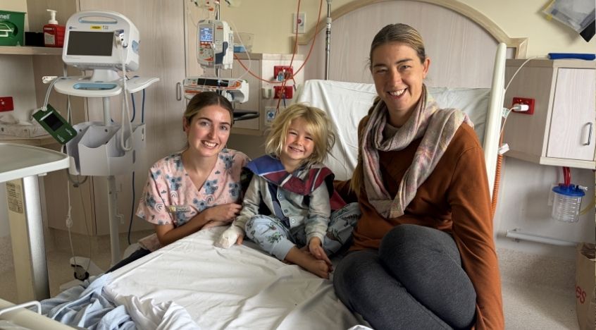A mother and child sit smiling on a hospital bed with a nurse beside them in the Gladstone Hospital Women and Family Centre.