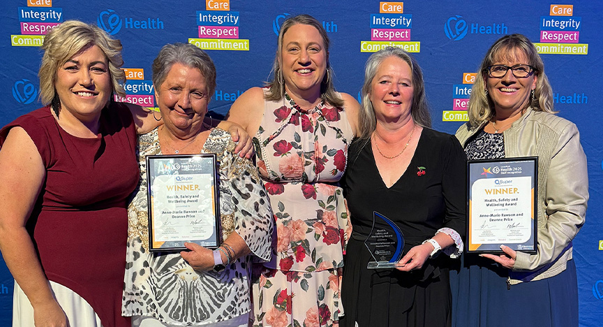 Winners of the Health, Safety and Wellbeing Award from Gladstone Hospital Pharmacy Department Anne-Marie Rawson (second left), and Deanna Price (right), are congratulated by Chief Executive Lisa Blackler, General Manager Gladstone and Banana Anita Pierantozzi, and Gladstone Hospital Director of Pharmacy Samantha Skilling. Healthcare workers wearing cocktail attire stand in front of a royal blue media wall printed with Central Queensland Health values at an awards presentation evening at a conference centre.