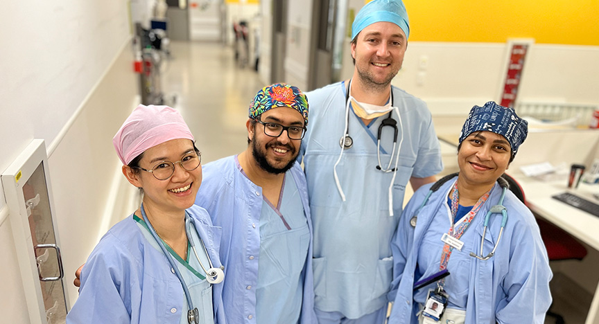 Four anaesthetic doctors wearing sky blue scrubs and scrub hats of different colours stand close together in the hallway connecting hospital operating theatres.