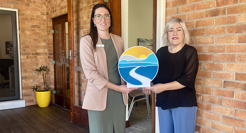 Tanya Johnson and Lisa Blackler stand side by side smiling at camera. Together, they're holding a sign of the Fitzroy Community Hospice logo. 