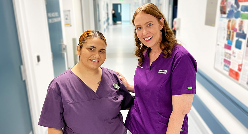 Two midwives wearing purple scrubs stand together in a hallway in a hospital maternity unit.