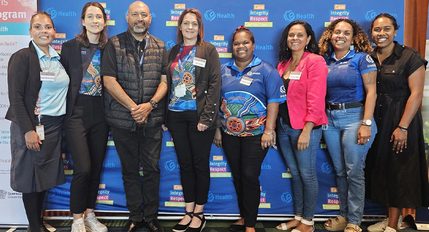 Group of First Nations health workers stand in front of a royal blue media wall with Central Queensland Health values and logos at a conference centre.